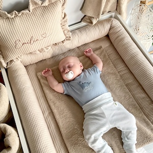 May include: A sleeping baby wearing a blue dinosaur-themed onesie and white pants rests on a beige waffle-textured changing pad. A matching pillow with the name "Emilia" embroidered on it and a ruffled edge is visible.