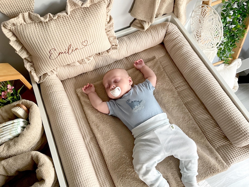 May include: A sleeping baby wearing a blue dinosaur-themed onesie and white pants rests on a beige waffle-textured changing pad. A matching pillow with the name "Emilia" embroidered on it and a ruffled edge is visible.
