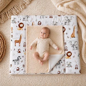 Può includere: Un bambino è sdraiato su un fasciatoio beige con una copertura bianca con una stampa di animali della savana. La stampa include leoni, giraffe, elefanti e zebre. Un asciugamano beige è posto sopra.