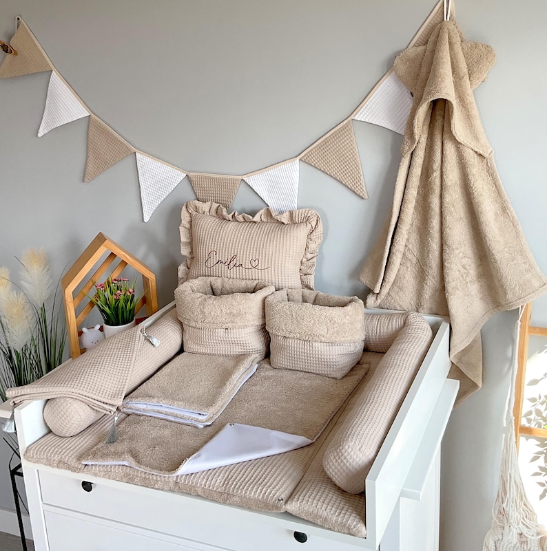 May include: A beige baby changing station set with a matching towel, pillow, and storage baskets. The set includes a changing pad, two bolsters, and a decorative banner. The pillow is embroidered with the name "Emilia".