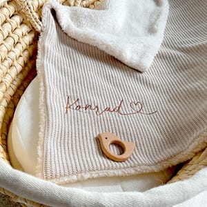 May include: A beige baby blanket embroidered with the name "Konrad" and a heart symbol in brown thread. A wooden bird-shaped teether rests on the blanket. The blanket is in a woven basket.