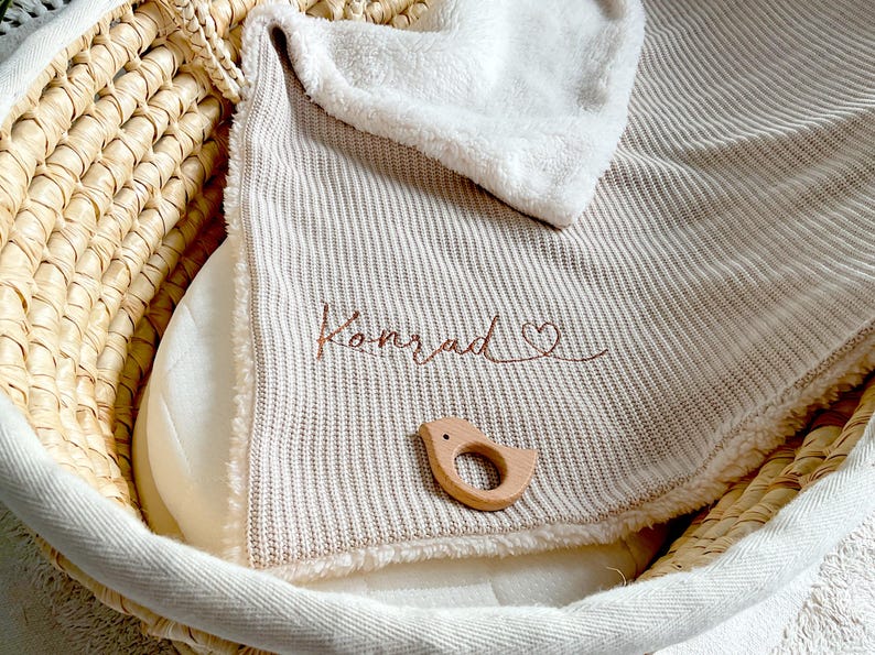 May include: A beige baby blanket embroidered with the name "Konrad" and a heart symbol in brown thread. A wooden bird-shaped teether rests on the blanket. The blanket is in a woven basket.