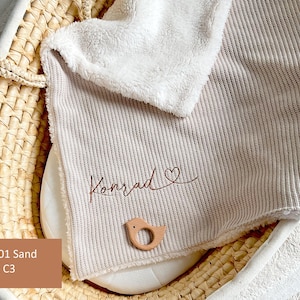 May include: A sand-coloured baby blanket with a ribbed texture, personalised with the name "Konrad" in a cursive font and a heart symbol. A small, wooden bird-shaped teether rests on the blanket. A white, fluffy blanket edge is visible.