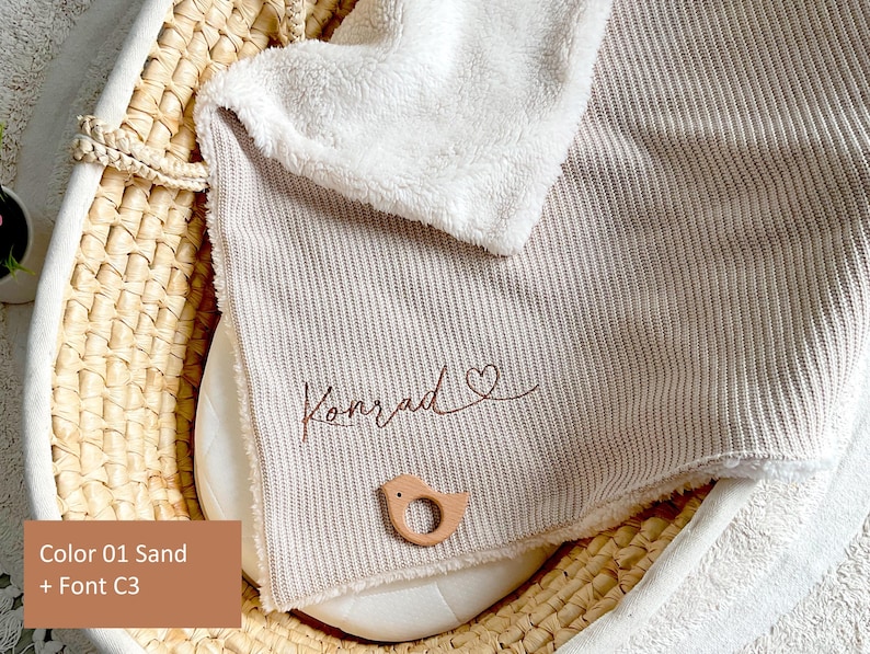 May include: A sand-coloured baby blanket with a ribbed texture, personalised with the name "Konrad" in a cursive font and a heart symbol. A small, wooden bird-shaped teether rests on the blanket. A white, fluffy blanket edge is visible.