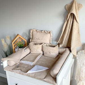 May include: A beige baby changing station set with a changing pad, pillows, and storage baskets. A matching hooded towel hangs on the wall. The set includes a pillow with the name "Emilia" embroidered on it.