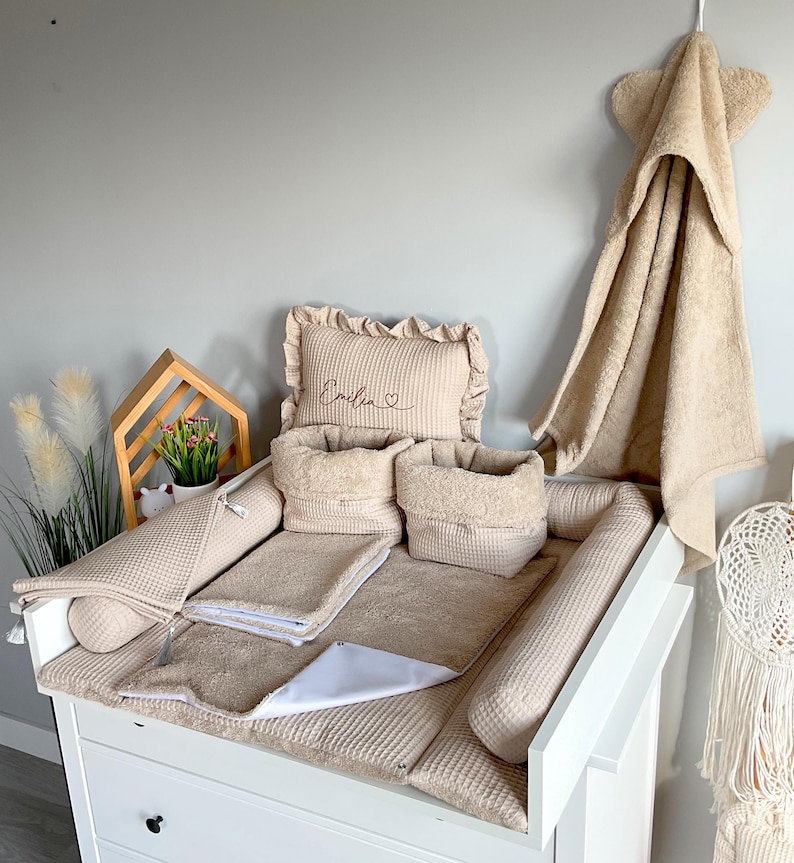 May include: A beige baby changing station set with a changing pad, pillows, and storage baskets. A matching hooded towel hangs on the wall. The set includes a pillow with the name "Emilia" embroidered on it.