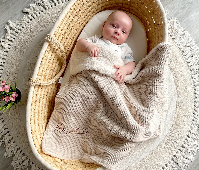 May include: A baby lies in a woven bassinet, covered with a beige blanket. The blanket is embroidered with the name "Konrad" and a heart. The bassinet sits on a round, fringed rug. A small pot of pink flowers is in the lower left corner.
