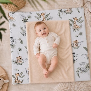 Puede incluir: Un bebé con un body color crema está acostado sobre un cambiador beige. El cambiador está sobre una alfombra blanca con estampado de animales de la jungla, incluyendo jirafas, cebras y tigres.