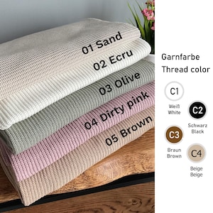 May include: A stack of five folded knitted blankets in colours labelled 01 Sand, 02 Ecru, 03 Olive, 04 Dirty pink, and 05 Brown. To the right, a chart displays thread colours labelled C1 White, C2 Black, C3 Brown, and C4 Beige with circular icons.