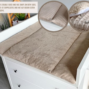May include: A beige, double-sided changing mat with snaps, placed on a white changing table. The mat has a textured surface and is designed for baby care. The text on the image reads: "THE MAT IS DOUBLE-SIDED AND HAS SNAPS ON BOTH SIDES."