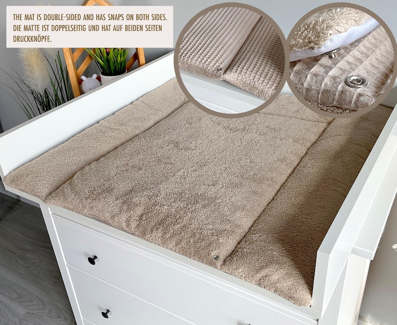May include: A beige, double-sided changing mat with snaps, placed on a white changing table. The mat has a textured surface and is designed for baby care. The text on the image reads: "THE MAT IS DOUBLE-SIDED AND HAS SNAPS ON BOTH SIDES."