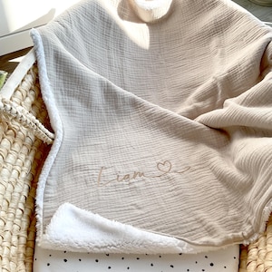 May include: A beige muslin baby blanket with a white sherpa lining. The blanket has the name "Liam" embroidered on it with a small heart.