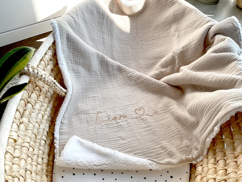 May include: A beige muslin baby blanket with a white sherpa lining. The blanket has the name "Liam" embroidered on it with a small heart.