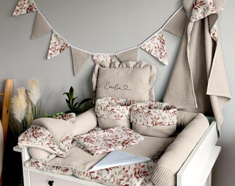 May include: A beige changing table with a white changing pad and a white and brown patterned border. The changing pad is decorated with a white and brown patterned pillow, two small baskets with a white and brown patterned design, and a larger pillow with the name "Emilia" embroidered on it. A white and brown patterned bunting hangs above the changing table.