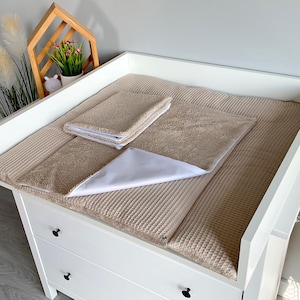 May include: A white changing table with a beige waffle-textured changing pad and matching towels. A white triangular cloth is placed on the pad. The changing table has three drawers with black knobs.