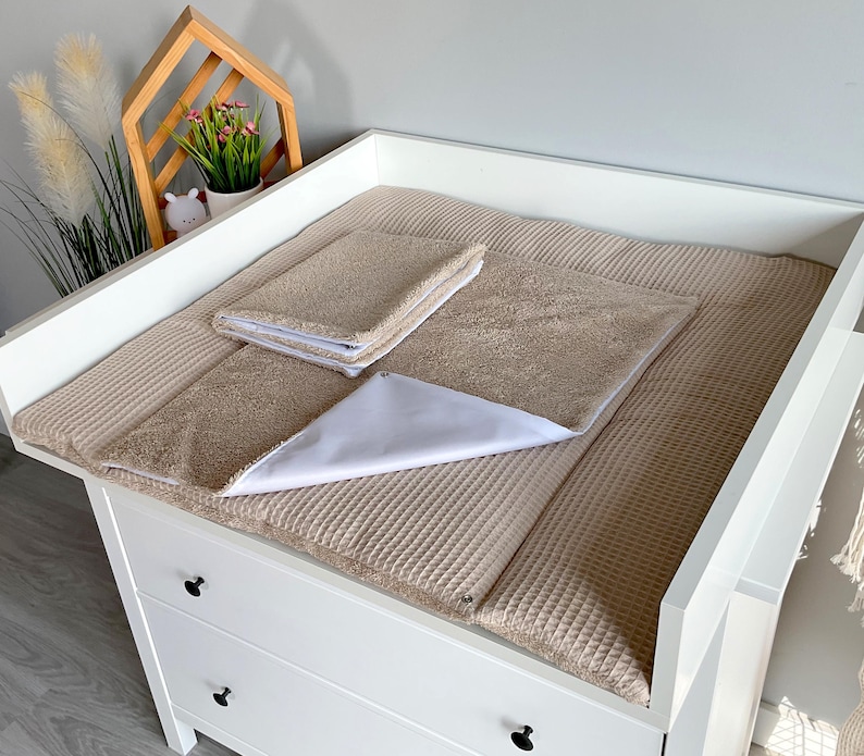 May include: A white changing table with a beige waffle-textured changing pad and matching towels. A white triangular cloth is placed on the pad. The changing table has three drawers with black knobs.