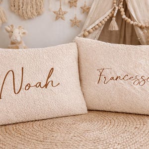 May include: Two beige rectangular cushions with embroidered names, "Noah" and "Francesca", in a brown script font. The cushions have a textured, plush appearance. The background includes a woven rug and decorative elements.