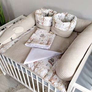 Peut inclure: Un lit bébé blanc avec un ensemble de literie à motifs beige et blanc. La literie comprend un pare-chocs, un drap-housse, une couverture et deux petits paniers. Le lit est décoré d'un animal en peluche en forme de renard.