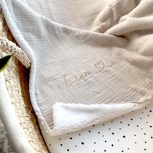 May include: A beige muslin baby blanket with a white sherpa lining. The blanket has the name "Liam" embroidered on it with a small heart.