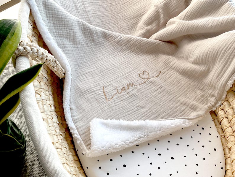 May include: A beige muslin baby blanket with a white sherpa lining. The blanket has the name "Liam" embroidered on it with a small heart.