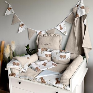 Peut inclure: Une table à langer blanche avec un matelas à langer en tissu gaufré beige et une serviette à capuche en tissu gaufré beige. Le matelas à langer est décoré d'un tissu blanc et beige avec un imprimé ours. La table à langer a également un coussin beige avec le nom "Emilia" brodé dessus. Il y a aussi plusieurs paniers beiges avec des imprimés ours.