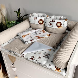 Könnte beinhalten: Ein weißer Wickeltisch mit einer beige strukturierten Wickelauflage und Stoßfängern. Das Set enthält zwei Stoffkörbe mit Löwen- und Affenaufdrucken, eine gemusterte Decke und ein hellblaues Tuch.