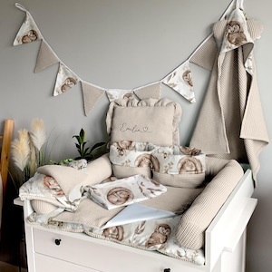Könnte beinhalten: Ein weißer Wickeltisch mit einer beige gepolsterten Auskleidung und einer Vielzahl von beige und weißen Babybettwäsche, darunter ein Kissen mit dem Namen "Emilia" bestickt. Die Bettwäsche zeigt einen Waldtierdruck mit Eulen und Eichhörnchen.