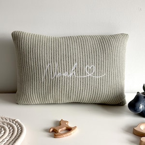 May include: A rectangular sage green knit cushion featuring the name Noah embroidered in white cursive script, followed by a small white embroidered heart outline. The cushion has a ribbed texture and is positioned horizontally on a flat surface.