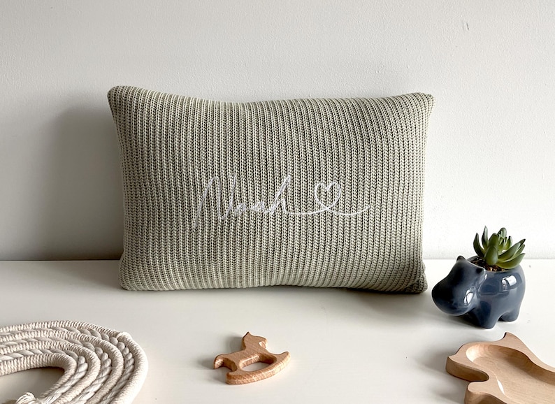 May include: A rectangular sage green knit cushion featuring the name Noah embroidered in white cursive script, followed by a small white embroidered heart outline. The cushion has a ribbed texture and is positioned horizontally on a flat surface.