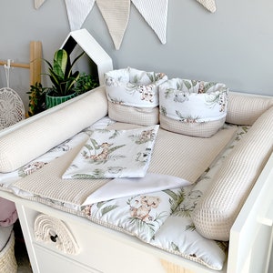 Könnte beinhalten: Ein weißer Wickeltisch mit einer neutralfarbenen, waffelstrukturierten Wickelauflage, Stoßfängern und zwei passenden Aufbewahrungskörben. Die Wickelauflage und die Körbe sind mit einem Dschungeltier-Print versehen. Eine weiße Decke und Kissen vervollständigen das Set.