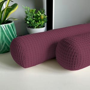 Op de afbeelding: Twee cilindrische kussens in bordeauxrood met een wafelstructuur. De kussens liggen op een wit oppervlak.