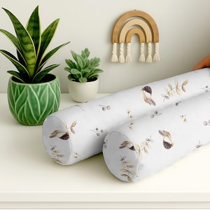 Op de afbeelding: Twee witte bolsterkussens met een vogel- en botanische print. De cilindrische kussens hebben een herhalend patroon van vogels, bladeren en bessen in gedempte tinten. De kussens liggen op een wit oppervlak.