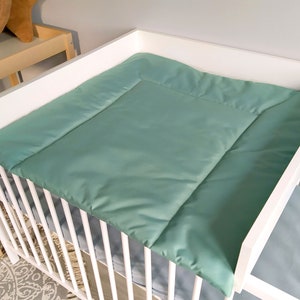 Op de afbeelding: Een witte commode met een lichtgroene verschoningsmat. De verschoningsmat is gemaakt van een zachte, gewatteerde stof. De commode staat in een babykamer.