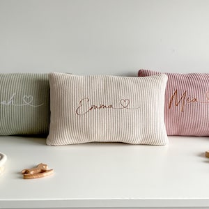 May include: Three rectangular knit cushions in sage green, cream, and dusty rose. Each cushion features embroidered cursive text and a small heart symbol. The left cushion reads Noah, the centre reads Emma, and the right cushion reads Mia.