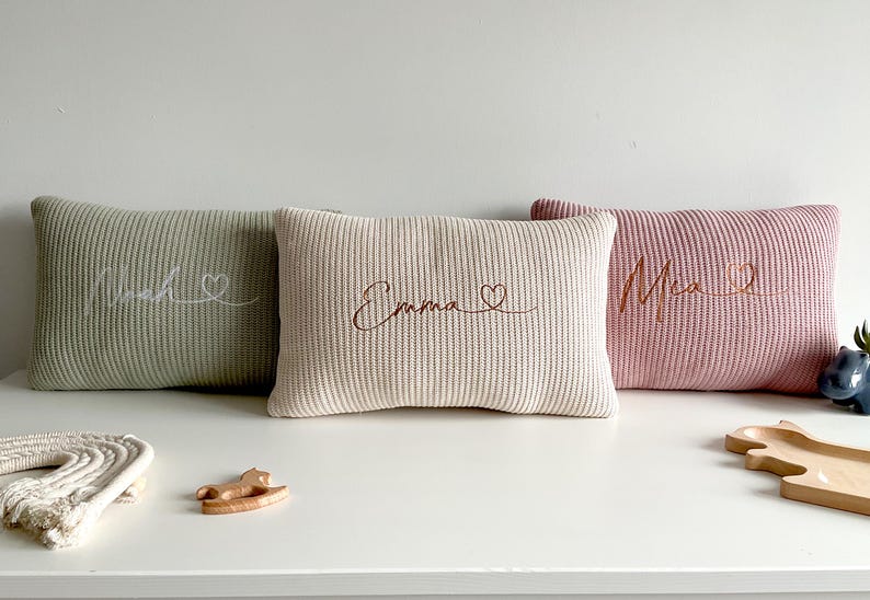May include: Three rectangular knit cushions in sage green, cream, and dusty rose. Each cushion features embroidered cursive text and a small heart symbol. The left cushion reads Noah, the centre reads Emma, and the right cushion reads Mia.