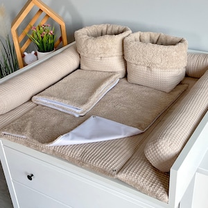 May include: A beige baby changing station with a white changing table. The station includes a changing pad, two cylindrical bumpers, two baskets, and a small towel. The changing pad and bumpers have a waffle texture.