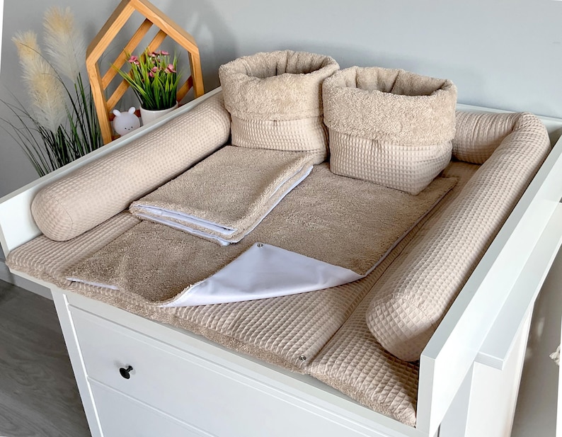 May include: A beige baby changing station with a white changing table. The station includes a changing pad, two cylindrical bumpers, two baskets, and a small towel. The changing pad and bumpers have a waffle texture.