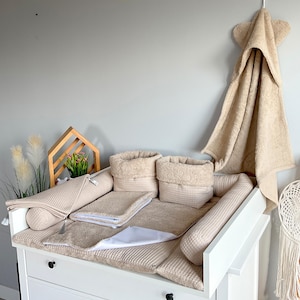 May include: A white changing table with beige waffle-textured accessories, including a changing pad, two storage baskets, and bolster pillows. A matching hooded towel hangs on the wall. The set is designed for a nursery.