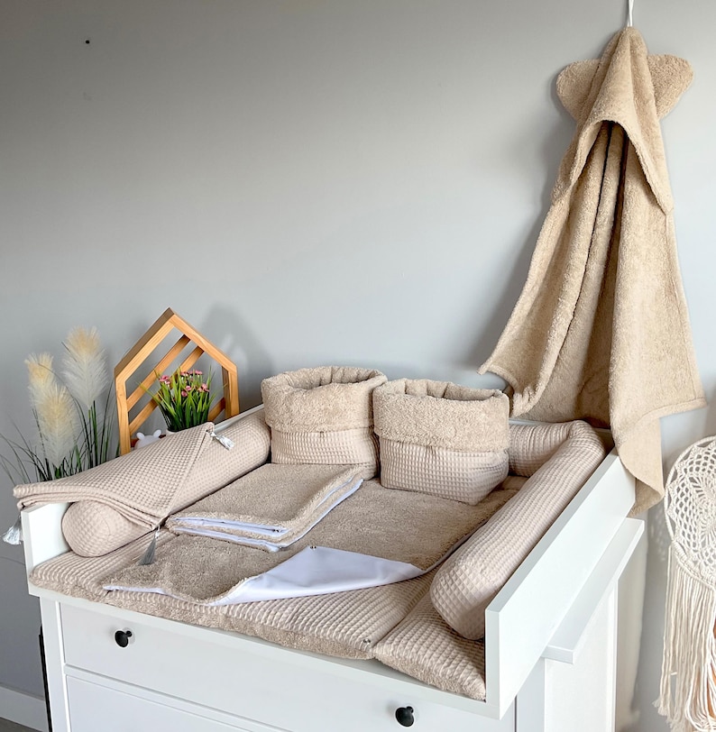 May include: A white changing table with beige waffle-textured accessories, including a changing pad, two storage baskets, and bolster pillows. A matching hooded towel hangs on the wall. The set is designed for a nursery.