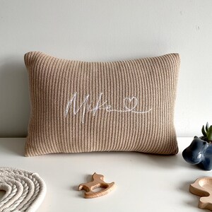 May include: A rectangular beige knit cushion featuring the embroidered white cursive text "Mike" followed by a small heart outline. The cushion has a ribbed texture and is positioned alongside a small blue hippo planter and wooden toys.