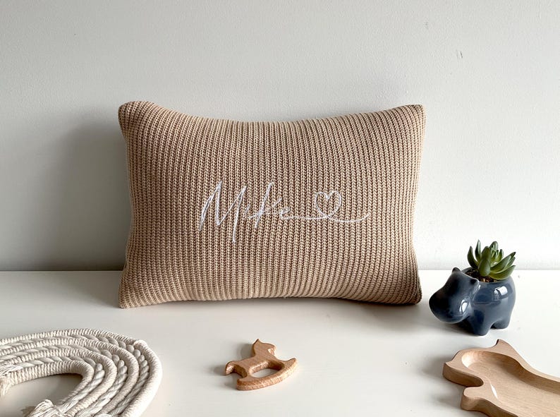 May include: A rectangular beige knit cushion featuring the embroidered white cursive text "Mike" followed by a small heart outline. The cushion has a ribbed texture and is positioned alongside a small blue hippo planter and wooden toys.
