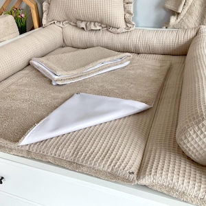 Può includere: Un fasciatoio beige con un cuscino a trama waffle, cuscini e un piccolo cuscino con bordo a balze. È visibile una fodera bianca per il fasciatoio. Il fasciatoio è su un comò bianco.