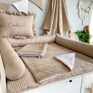 Può includere: Un fasciatoio beige con un motivo a nido d'ape. Include un cuscino con il nome "Emilia" ricamato, un fasciatoio e due cuscini cilindrici. Un asciugamano e un festone coordinati sono sullo sfondo.