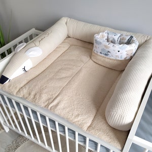 Op de afbeelding: Een wit wiegje met een beige gevulde bumper en een wit, pluche, dier-vormig kussen. De bumper heeft een wit, gestikt ontwerp en het kussen heeft het woord "Eska" geborduurd. Er is een kleine, beige, gevulde mand met een blauwe en witte gepatroneerde voering.