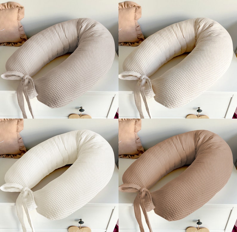 May include: Four waffle-textured pregnancy pillows in beige, white, and brown. The pillows are shaped like a horseshoe and have a tied ribbon at the opening.