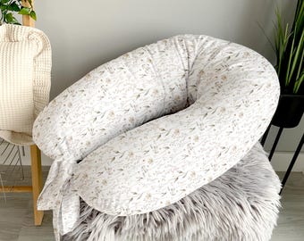 May include: A white and beige floral print pregnancy pillow with a U-shape design. The pillow is on a gray fluffy surface.