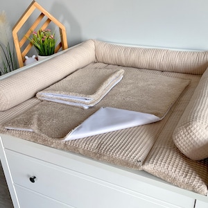 May include: A beige changing table with a padded surface and raised sides. It includes a matching towel and a white changing pad. The table is set against a light-colored wall, with decorative elements in the background.