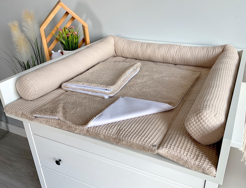 May include: A beige changing table with a padded surface and raised sides. It includes a matching towel and a white changing pad. The table is set against a light-colored wall, with decorative elements in the background.