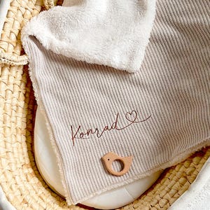 May include: A beige and white baby blanket with the name "Konrad" embroidered in brown script, along with a heart symbol. A wooden bird-shaped teether rests on the blanket, inside a woven basket.