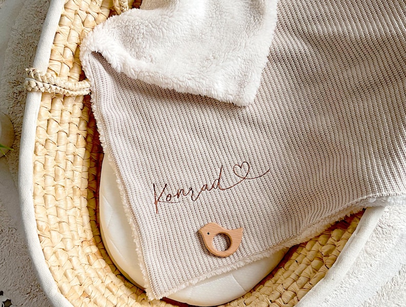 May include: A beige and white baby blanket with the name "Konrad" embroidered in brown script, along with a heart symbol. A wooden bird-shaped teether rests on the blanket, inside a woven basket.